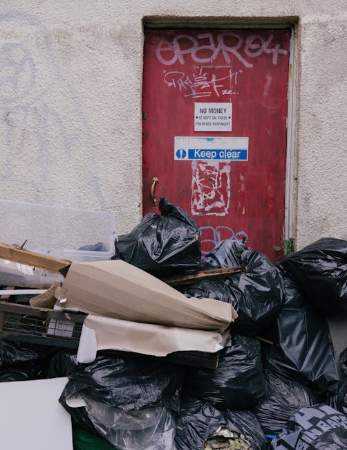 A collection of black refuse bags and scattered cardboard and packaging materials piled against a textured, off-white wall near a small, red metal door covered in graffiti and stickers. The bags are stacked haphazardly, with some leaning against the lower part of the doorframe and others spilling onto a concrete pavement. The door features various graffiti tags in white and pink spray paint, along with a small white sign stating 'No Money' and a blue and white sticker with the words 'Keep Clear'. Surrounding the rubbish, there are some loose cardboard pieces, a partially visible shopping cart, and minimal greenery growing near the base of the wall. The scene suggests an environment where independent waste collection might be carried out, aligning with private rubbish clearance services such as those provided by Waste Clearance Earls Court, and reflects common urban refuse accumulation near exterior building entrances.