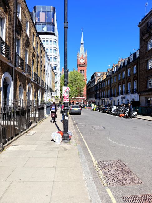 A wide urban street scene in a city environment during daytime, featuring a sidewalk on the left with several pedestrians walking, including a person standing near a small pile of white rubbish bags placed next to a black metal fence. The pavement appears clean and well-maintained. To the right, the street is lined with parked motorcycles and a few cars, some covered with tarps or protective covers. In the background, a prominent historic red-brick clock tower with a tall, pointed spire and a clock face is visible against a clear blue sky, contrasting with the modern high-rise building that has a curved glass facade further left. A central streetlight pole is visible in the foreground, slightly obstructing part of the view. The environment suggests a busy city area where private waste collection and rubbish clearance services, like those offered by Waste Clearance Earls Court, might operate to manage waste and rubbish removal efficiently in this urban setting.