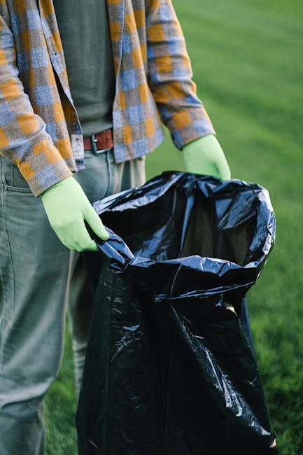 A person wearing a plaid shirt with yellow, gray, and blue tones, along with gray trousers, is holding open a large black trash bag lined with a silver inner layer, which appears to be made of plastic. The individual’s hands are gloved in bright green latex gloves, and they are standing outdoors on a grass surface with a blurred green background, suggesting a garden or park setting. The scene relates to waste collection or rubbish disposal, demonstrating a private or independent approach to rubbish removal activities, possibly by Waste Clearance Earls Court. The image focuses on the handling of waste materials, emphasizing environmentally conscious or on-site clearance practices, with natural lighting highlighting the textures of the gloves, clothing, and trash bag surface. This visual supports content related to rubbish removal costs by illustrating the process of rubbish collection and the preparation for disposal or recycling, typical of private waste management services. The environment appears clean and organized, aligning with professional waste clearance standards.