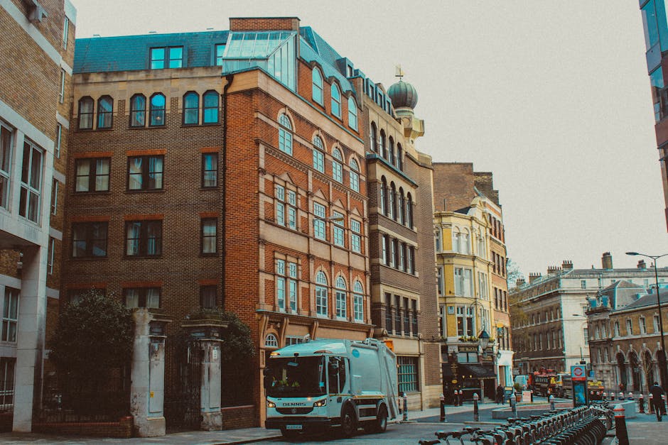 The image depicts a city street scene featuring a row of older, multi-storey buildings with distinct architectural details, including large arched and rectangular windows, some with decorative arches, and a mix of brick and stone facades. The prominent building in the center has a reddish-brown brick exterior with patterned brickwork and white framing around its windows, topped by a green copper roof with dormer windows. To its right, there is a cream-colored building with intricate mouldings, large bay windows, and a rounded turret-like corner, giving a Victorian-era appearance. In the foreground, a light blue waste collection truck labeled with the company name Waste Clearance Earls Court is parked on the street, close to a low perimeter wall with columns and wrought iron fencing, suggesting private property. The street is lined with black bollards and bicycle racks, and the scene is illuminated by natural daylight, capturing an urban environment prepared for or engaged in rubbish removal or waste collection, with a focus on the buildings, the waste truck, and surrounding city infrastructure.