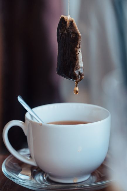 A close-up of a white ceramic cup filled with hot tea, placed on a saucer with a small metal spoon resting inside. Above the cup, a tea bag with dark, textured material is suspended by a string, with a small droplet of liquid hanging from its lower edge. The background is softly blurred with warm, natural lighting, emphasizing the focus on the cup and tea bag, and creating a calm, inviting atmosphere. This scene highlights the process of brewing or preparing hot beverages, with subtle references to waste disposal through the tea bag and spoon as part of everyday kitchen waste, relevant to rubbish removal and waste management services provided by Waste Clearance Earls Court.