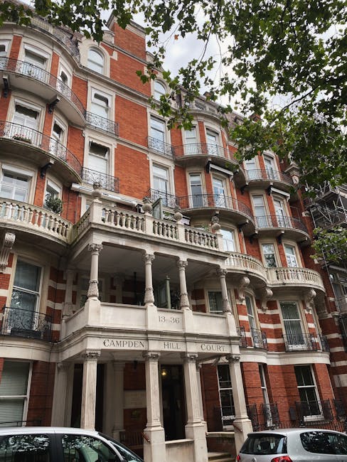 The image depicts a multi-storey residential building with a distinctive Victorian or Edwardian architectural style, featuring red brickwork with contrasting white stone or stucco details. The facade includes several prominent protruding bay windows with black metal railings, and the building's corners are rounded, enhancing its ornate appearance. The entrance is situated beneath a covered portico supported by classic columns, with the address number 13-36 and the name 'Campden Hill Court' inscribed on the front. Above the entrance, a decorative balcony with a balustrade extends from the second floor, and additional balconies with similar railings are visible on the upper floors. The scene is shaded by leafy green trees, their branches extending across the top of the image, creating a natural canopy. Parked cars are visible along the street in front of the building, and scaffolding can be seen on part of the right side, indicating ongoing maintenance or renovation work. The overall setting suggests an urban residential area where private or independent rubbish clearance might be necessary, aligning with services provided by Waste Clearance Earls Court, especially given the context of managing waste in such detailed building environments.