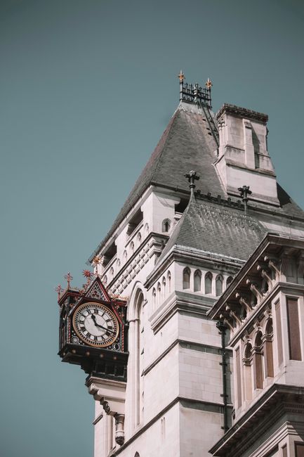 Close-up view of a historic clock tower on a white stone building, featuring ornate architectural details such as decorative cornices and arched windows. The clock face shows a time close to 2:15, with black roman numerals and hands against a white background, encased in a red and gold frame that extends outward from the building's facade. The tower has a steep, dark grey slate roof topped with decorative ironwork finials and a weather vane. The surrounding environment is minimal, with a clear, slightly overcast sky providing a neutral backdrop. This architectural feature is part of an older, multi-story structure, exemplifying traditional design elements often seen in British civic buildings. Such buildings may be involved in private or public functions, including activities related to waste management or service provision, as conceptualised by Waste Clearance Earls Court in the context of search relevance for rubbish and waste removal services in London.