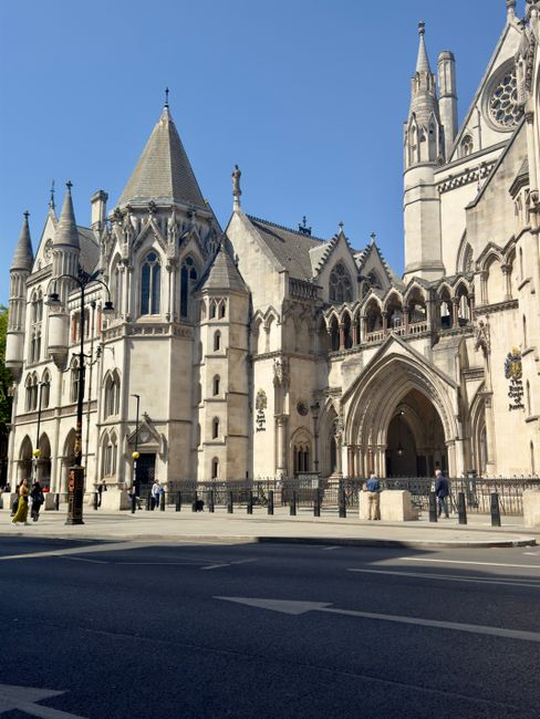The image depicts a historic Gothic-style cathedral with intricate architecture, including pointed arches, spires, and decorative stone carvings. The façade features large arched entrance doors and tall, narrow stained glass windows. The building is constructed from light-colored stone with detailed detailing along the buttresses and turrets. In the foreground, there is a paved street with a few pedestrians walking along the sidewalk, and a series of bollards line the edge of the pavement to prevent vehicle access. The scene is illuminated by natural daylight, casting minimal shadows and highlighting the textures of the stonework. Limited greenery is visible with a small tree at the left edge of the image. This setting suggests a busy urban area with vehicles, and the cathedral stands prominently as an architectural landmark. As part of the visible environment, it may be located near areas where private waste management services, such as those provided by Waste Clearance Earls Court, are used for maintenance and cleanup of surrounding urban spaces.